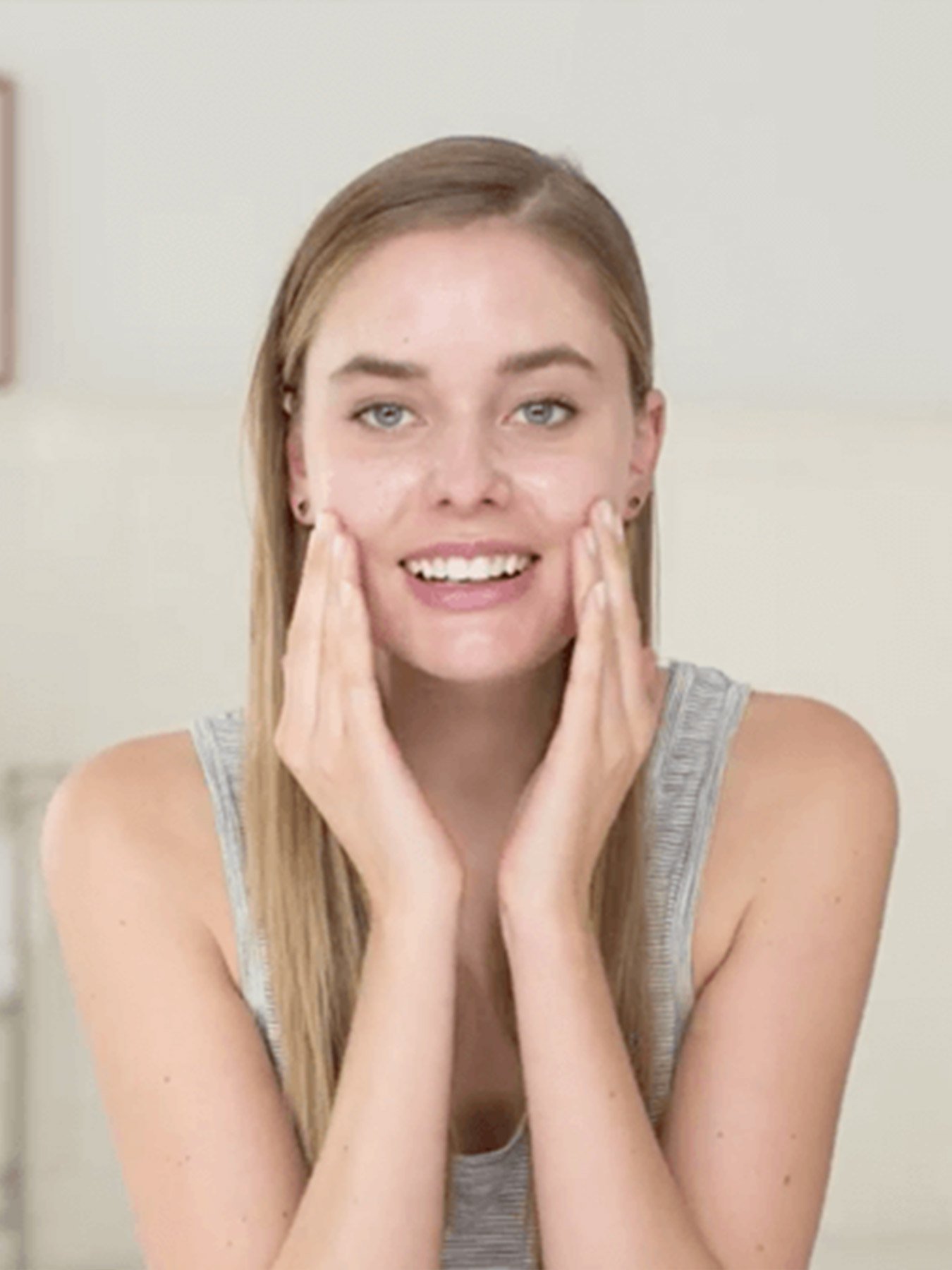 A woman wearing a grey tank top in a white room applying Clearly Brighter Argan Nut Gentle Exfoliating Cleanser to her face.
